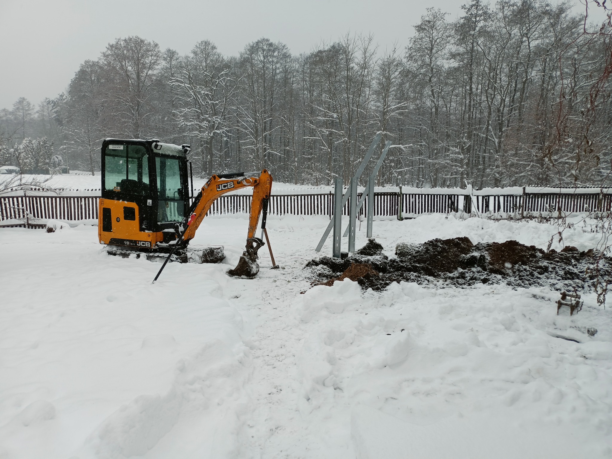 Żółta mini-koparka JCB na śniegu, obok wykopana ziemia i metalowe słupki, zimowy krajobraz w tle.