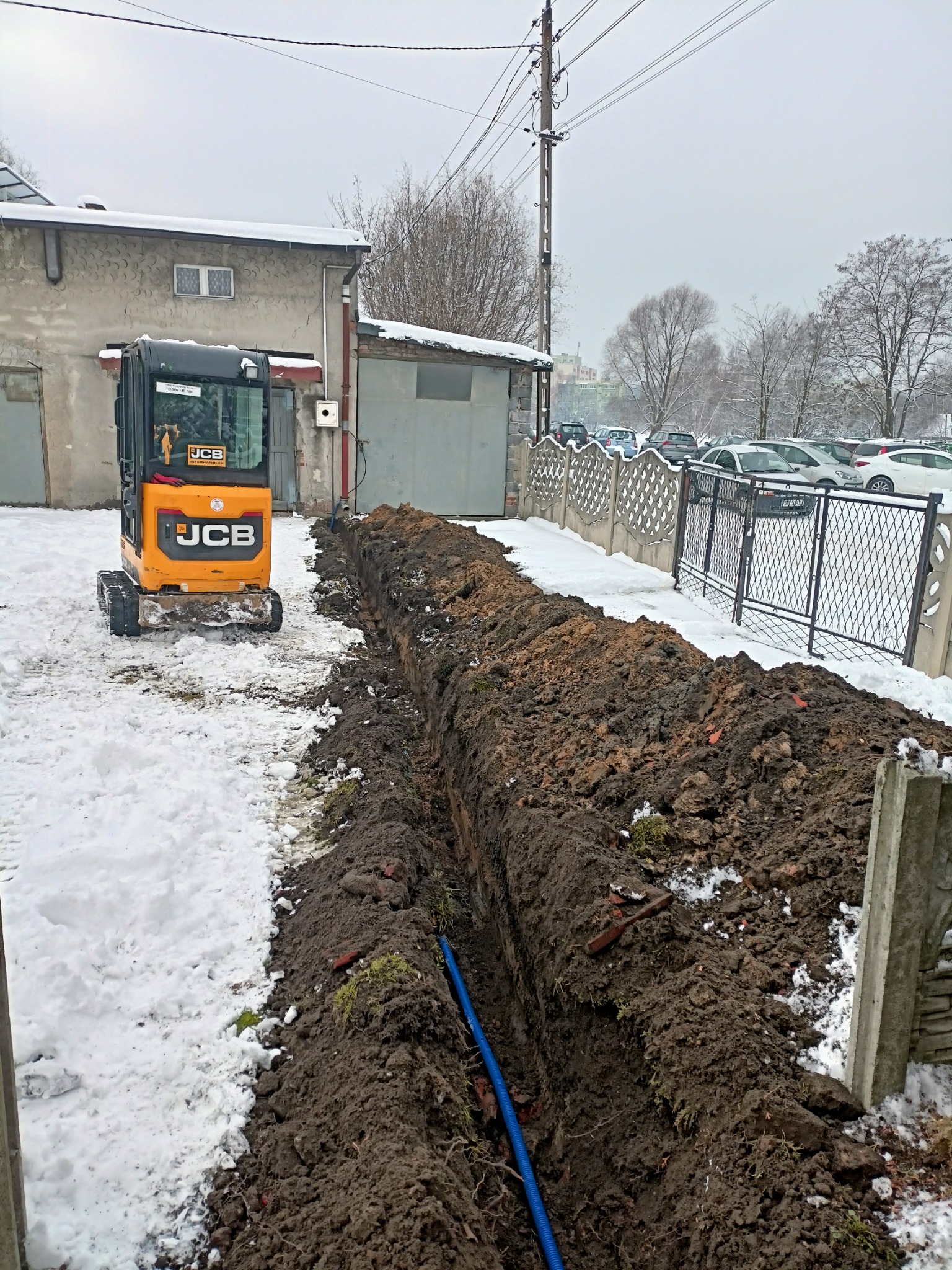 Żółta minikoparka JCB stoi obok wykopanego rowu z niebieską rurą w środku, w tle budynek i częściowo ośnieżony teren.