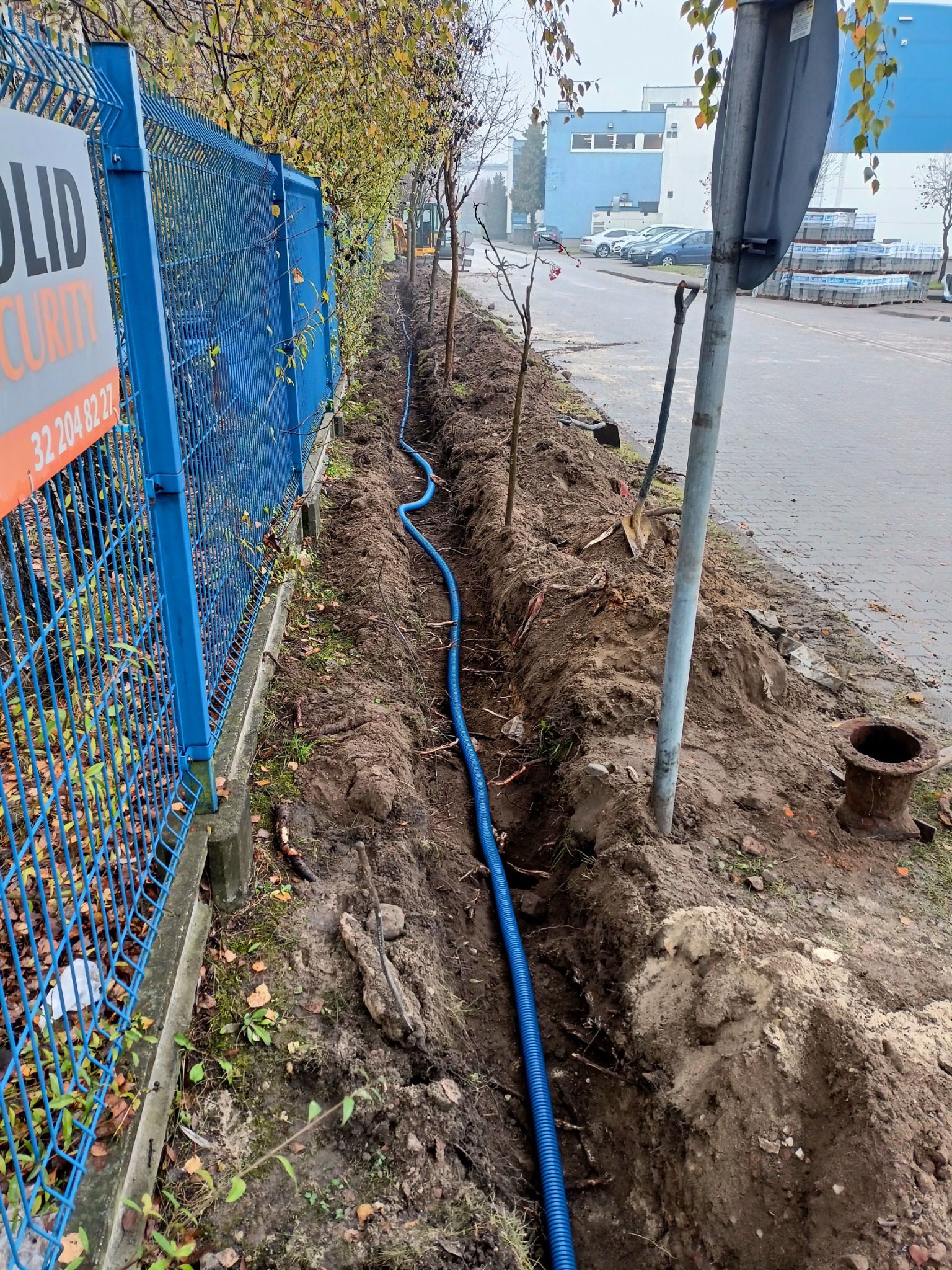Wąski wykop z niebieską rurą karbowaną ułożoną wzdłuż ogrodzenia z siatki, widoczne narzędzia, słupki i fragment ulicy z zaparkowanymi samochodami w tle.