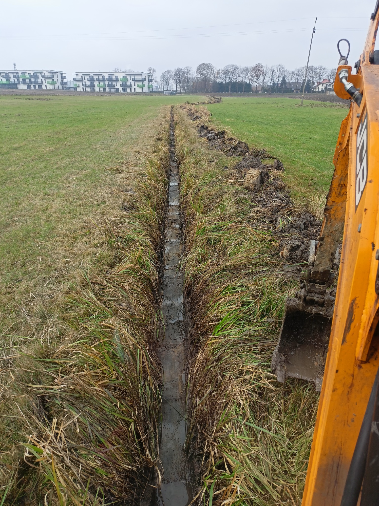 Wąski rów wypełniony wodą, wykopany na podmokłym terenie, widoczna łyżka koparki JCB po prawej stronie, w tle budynki mieszkalne.
