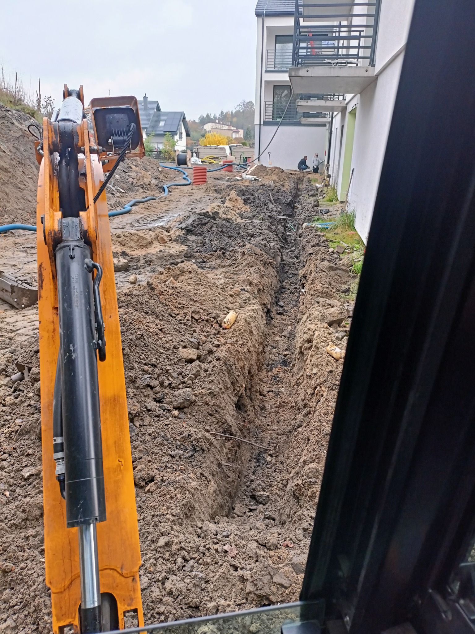 Wąski wykop w ziemi, widziany z kabiny pomarańczowej koparki; w tle budynek mieszkalny i pracownicy przy wykopie.