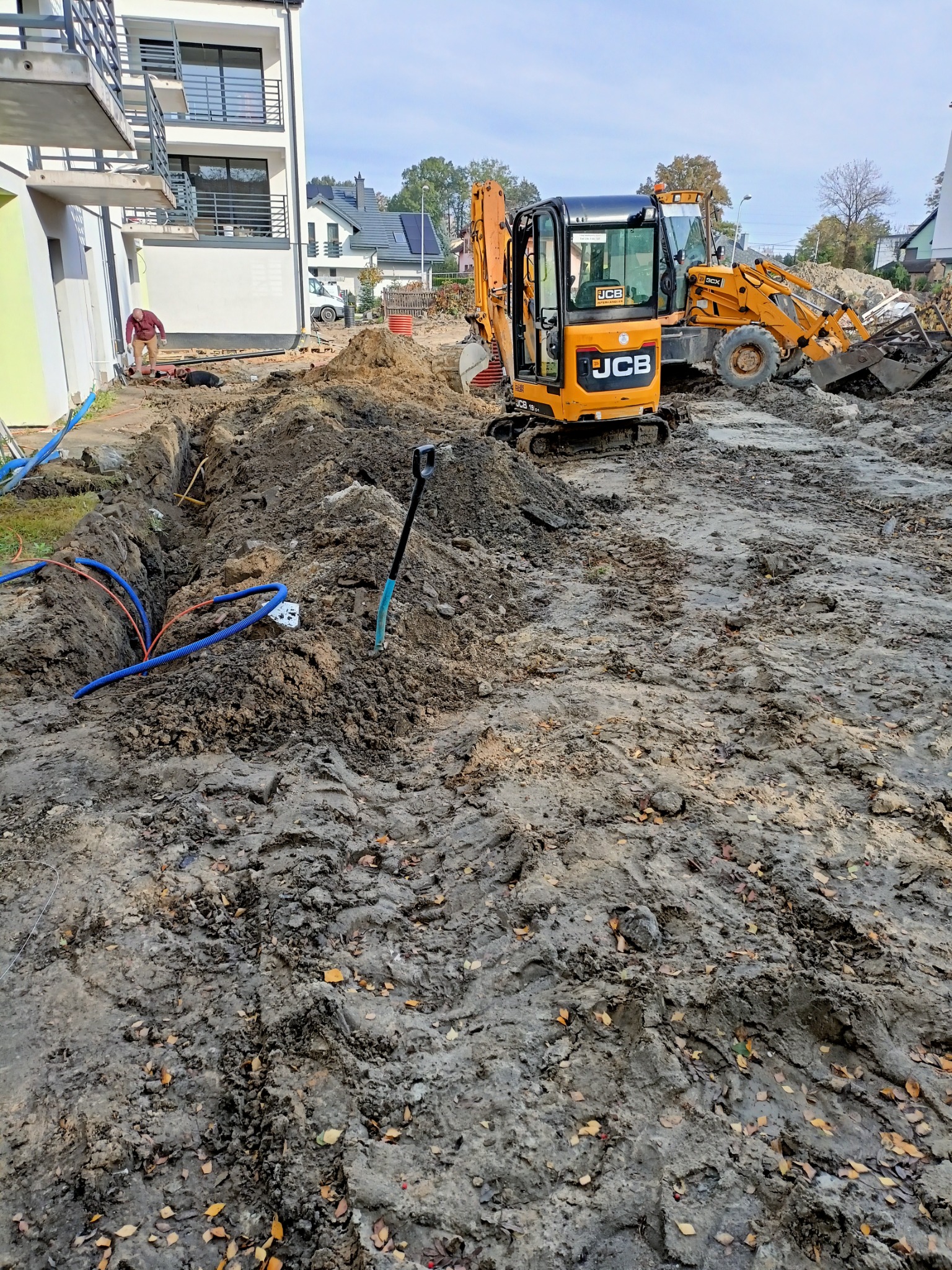 Żółty mini-koparka JCB i koparko-ładowarka na placu budowy obok wykopu z rurami, w tle budynek mieszkalny i pracownik.