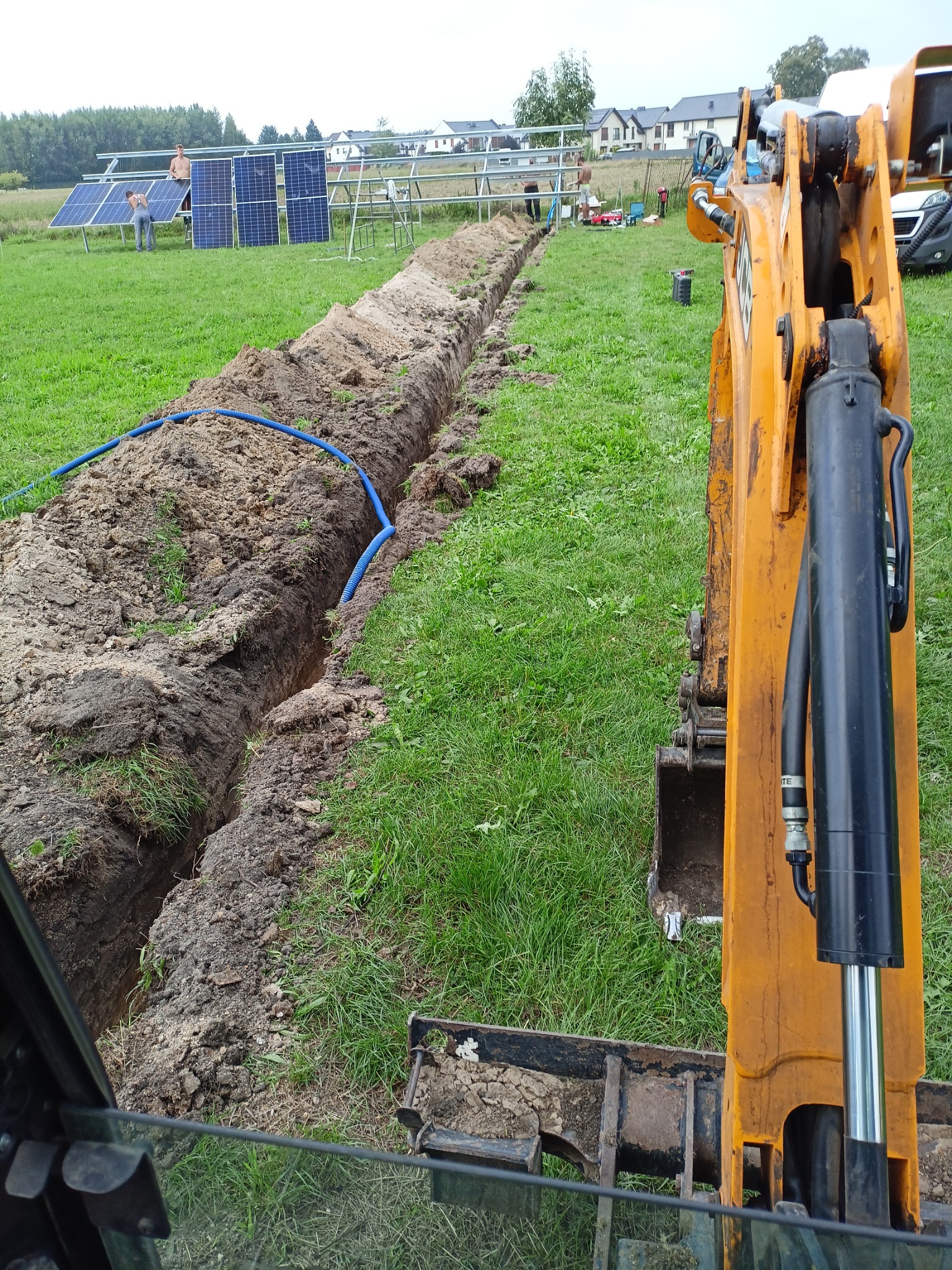 Wykopany wąski rów w trawiastym terenie z niebieską rurą w środku, z tyłu instalacja paneli słonecznych i zabudowa mieszkalna, widok z kabiny pomarańczowej koparki.