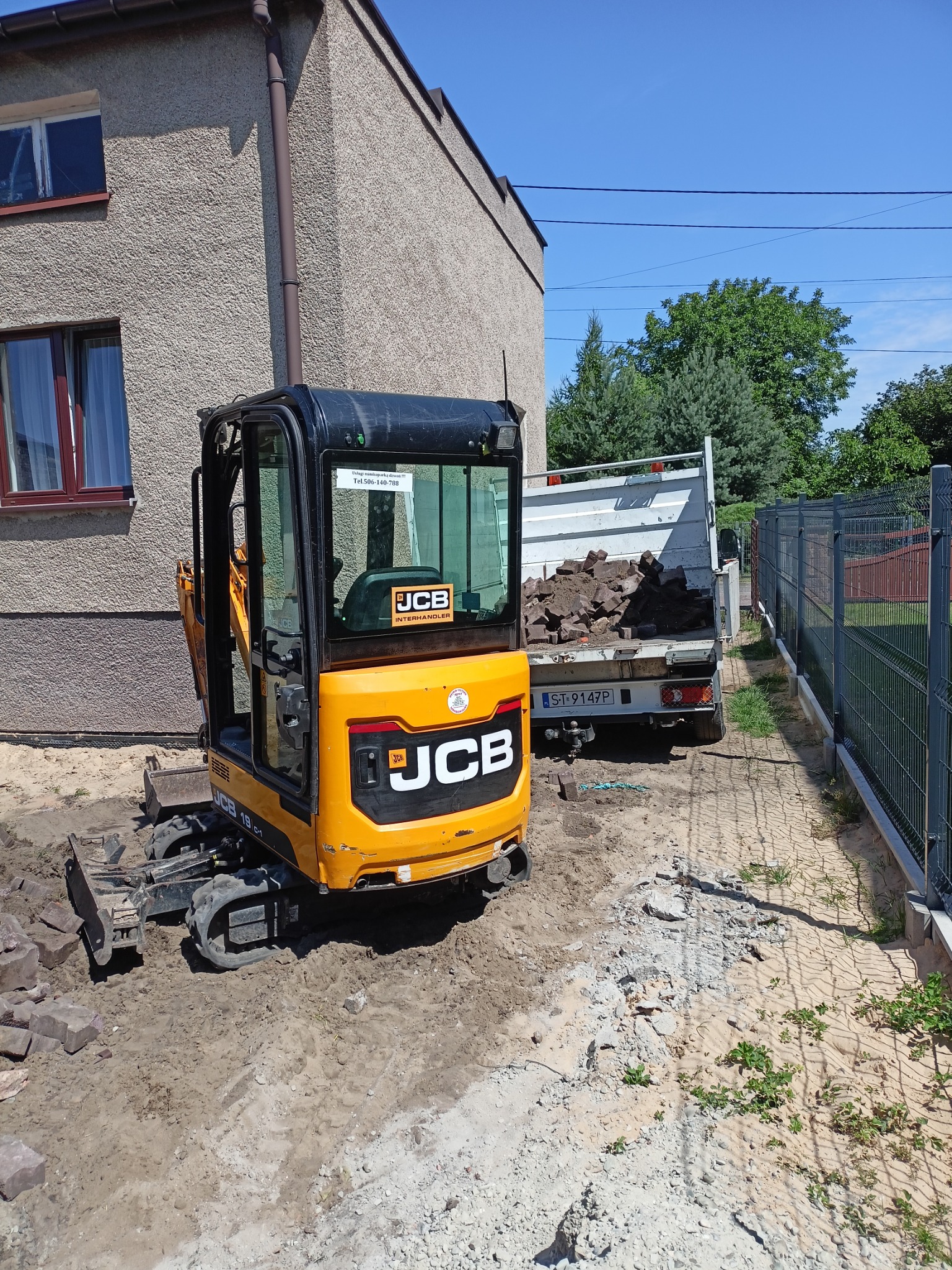 Żółta mini koparka JCB 19C-1 na placu budowy, obok zaparkowany samochód ciężarowy z ładunkiem ziemi i gruzu, w tle budynek mieszkalny i ogrodzenie.
