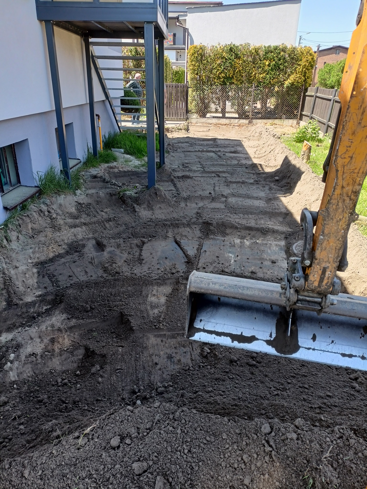 Żółta koparka z łyżką w trakcie wyrównywania terenu przy budynku z metalowymi schodami zewnętrznymi, widoczne świeże ślady opon i wykopana ziemia.