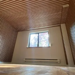 Pokój z drewnianymi listwami dekoracyjnymi na ścianach i suficie, tworzącymi geometryczny wzór. Widoczne okno z widokiem na drzewa, grzejnik pod oknem i drewniana podłoga.