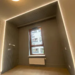 Pusta wnęka z oknem, grzejnikiem i oświetleniem LED, wykończona panelami ściennymi i drewnianą podłogą w jodełkę.