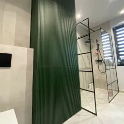 Nowoczesna łazienka z kabiną prysznicową typu walk-in z czarnymi profilami i zieloną, pionowo żłobioną ścianką działową.