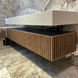 Nowoczesna łazienka z wiszącą szafką umywalkową z lamelami z jasnego drewna i blatem z białego konglomeratu na tle marmurowych płytek.