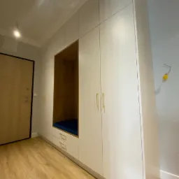 Biała zabudowa meblowa z wnęką do siedzenia z granatową poduszką, złotymi uchwytami i drewnianą podłogą w przedpokoju.