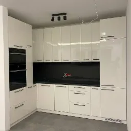 Nowoczesna, biała kuchnia z czarnym blatem i matowymi, poziomymi uchwytami, oświetlona trzypunktową lampą sufitową, z widocznym kablem wystającym z sufitu.