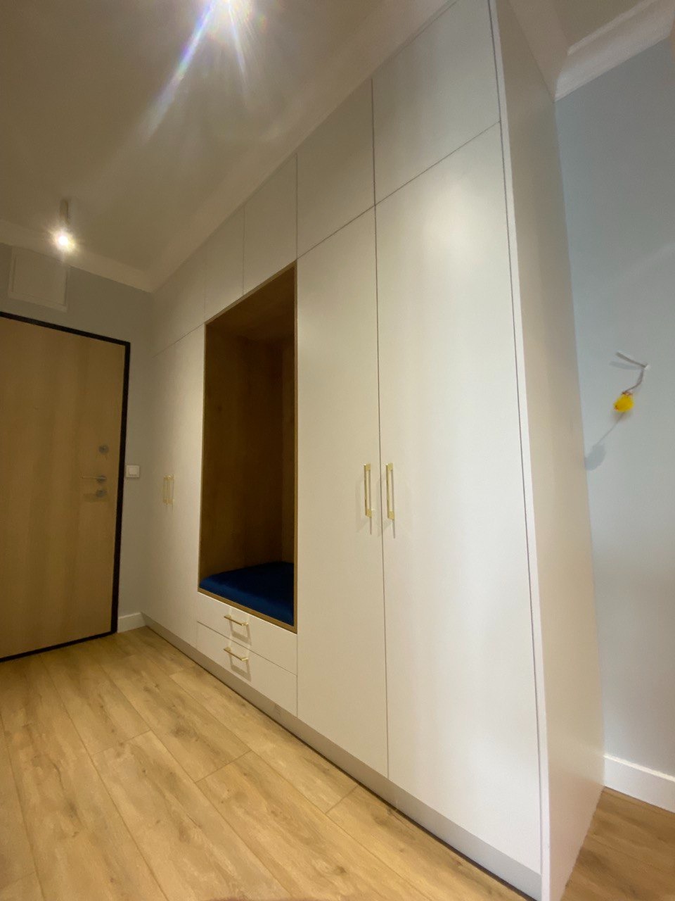 Biała zabudowa meblowa z wnęką do siedzenia z granatową poduszką, złotymi uchwytami i drewnianą podłogą w przedpokoju.