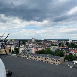 Antena satelitarna i antena naziemna na dachu budynku z widokiem na panoramę miasta pod pochmurnym niebem.