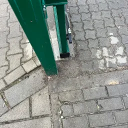 Fragment zielonej, metalowej konstrukcji bramy na tle szarej kostki brukowej. Widoczne ślady użytkowania i zabrudzenia na metalu i kostce.