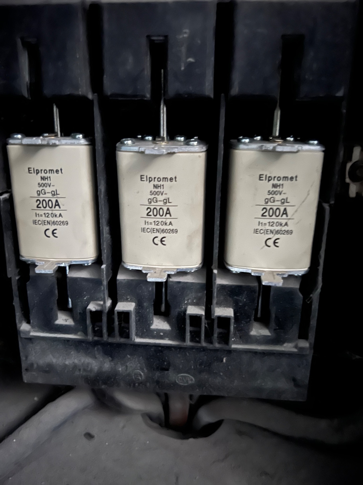 Trzy bezpieczniki topikowe Elpromet NH1 200A w czarnej, brudnej obudowie rozdzielnicy elektrycznej, widoczne oznaczenia napięcia i prądu.