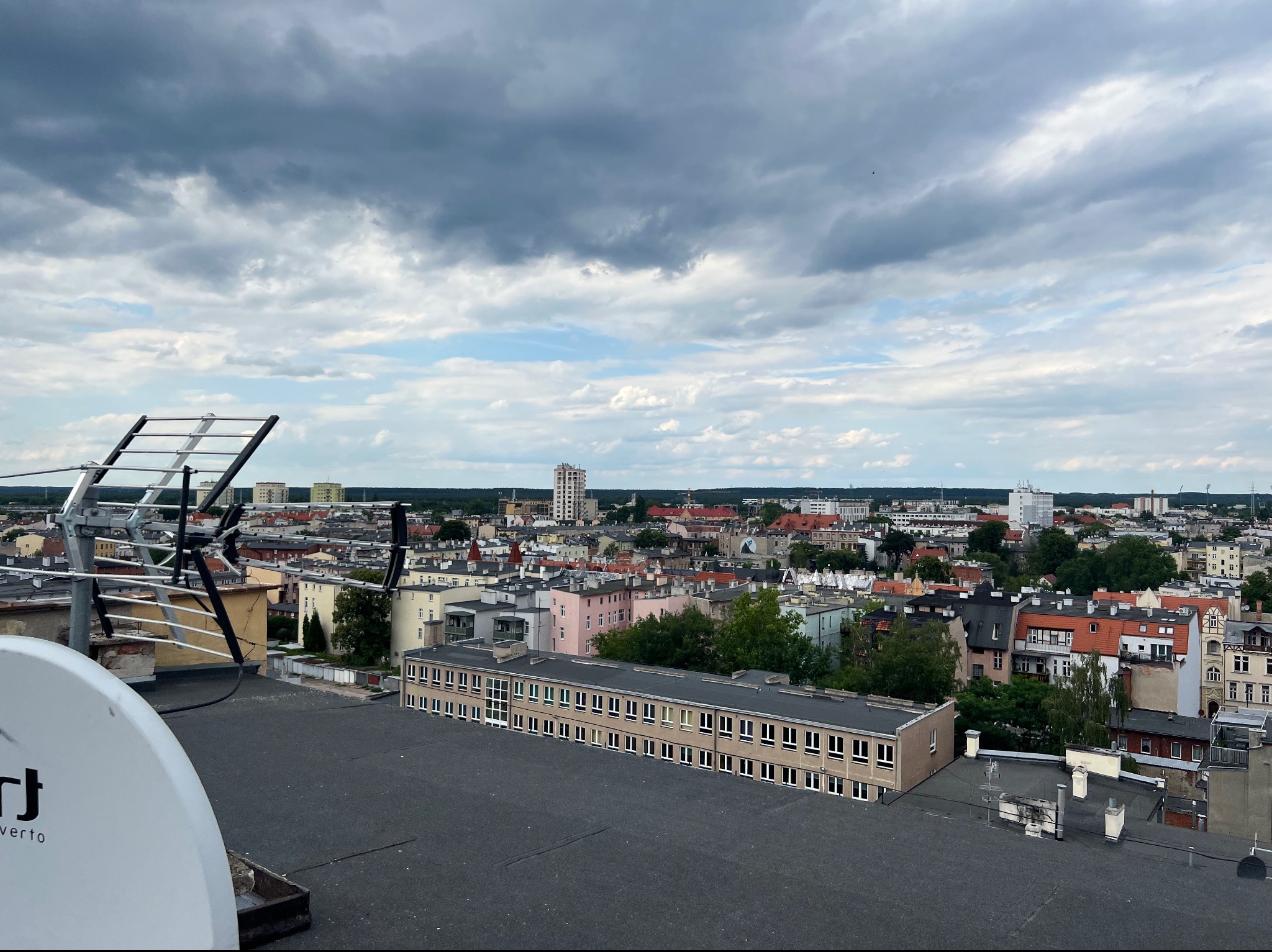 Antena satelitarna i antena naziemna na dachu budynku z widokiem na panoramę miasta pod pochmurnym niebem.