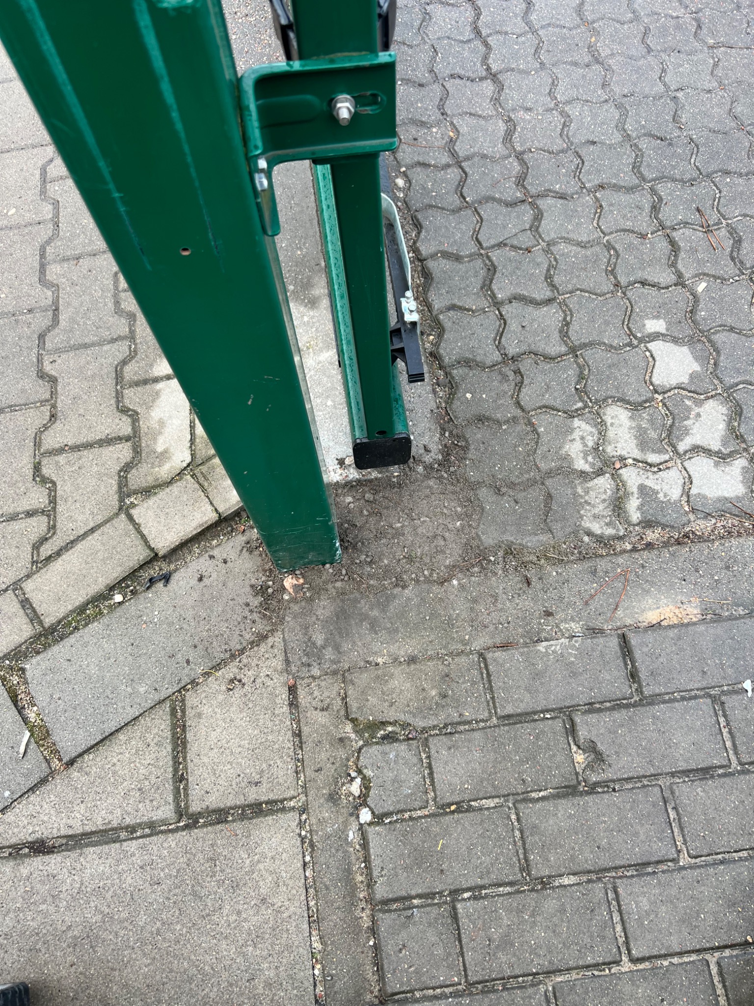 Fragment zielonej, metalowej konstrukcji bramy na tle szarej kostki brukowej. Widoczne ślady użytkowania i zabrudzenia na metalu i kostce.