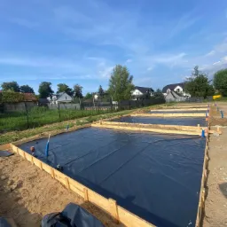 Przygotowane szalunki z folią pod wylewkę fundamentów na piaszczystym gruncie, widok na kilka działek w tle.