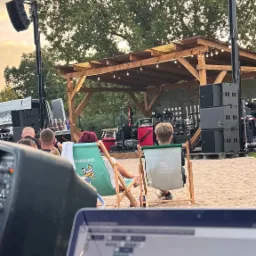 Plenerowa scena z nagłośnieniem, oświetleniem i publicznością siedzącą na leżakach. W tle widoczna scena z instrumentami. Laptop na pierwszym planie.