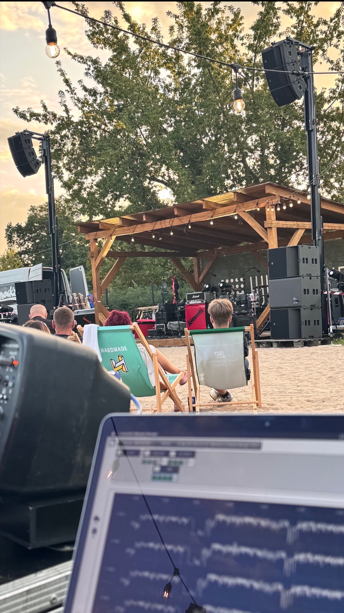 Plenerowa scena z nagłośnieniem, oświetleniem i publicznością siedzącą na leżakach. W tle widoczna scena z instrumentami. Laptop na pierwszym planie.