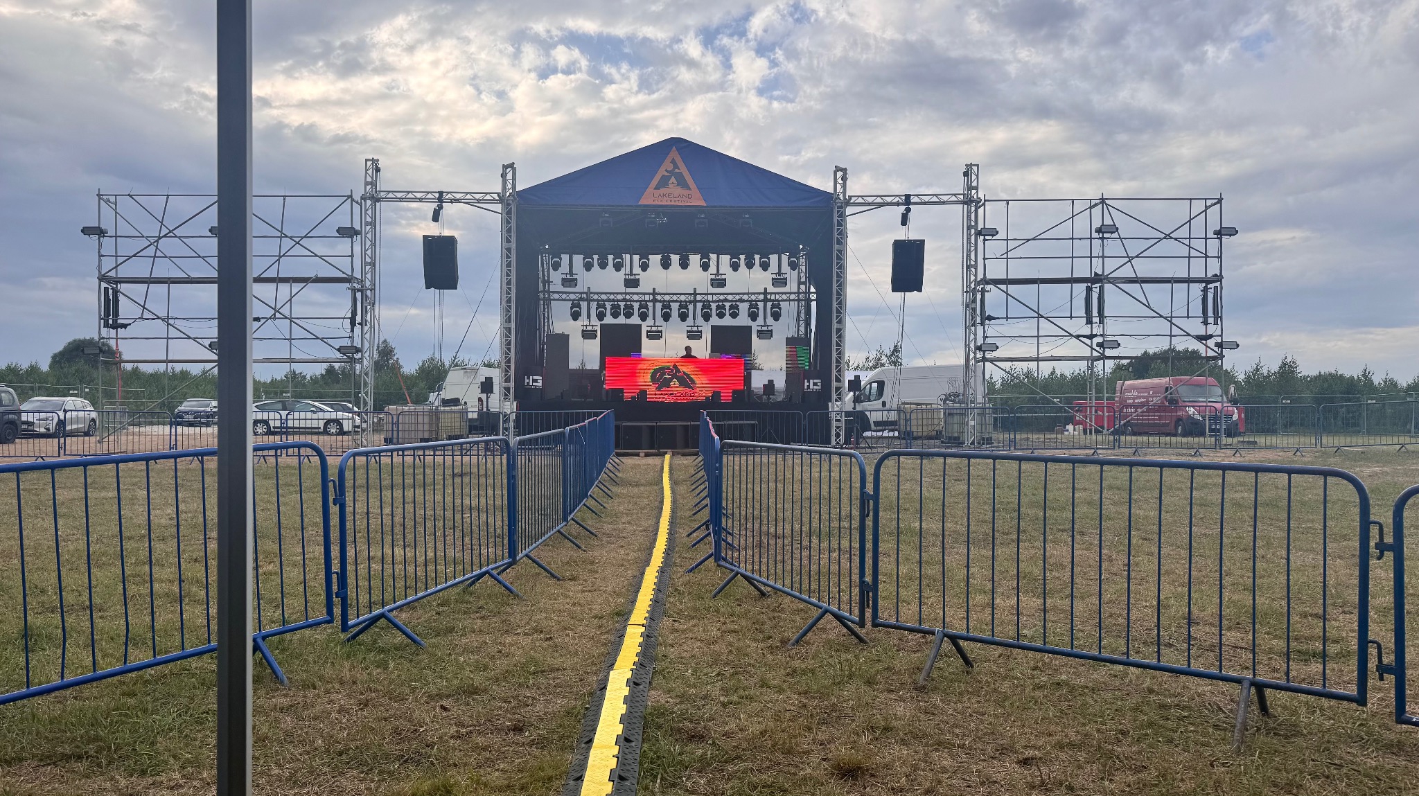 Scena plenerowa z nagłośnieniem i oświetleniem, przygotowana do koncertu. Widoczne barierki, scena z konstrukcją, ekrany LED i zaplecze techniczne.