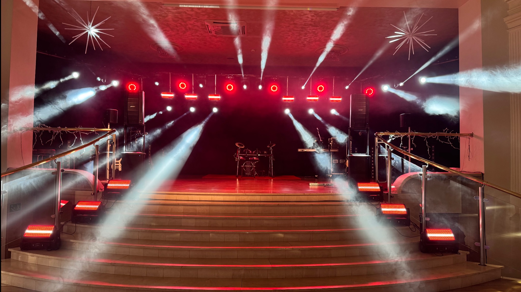 Oświetlona scena z instrumentami, reflektorami i efektem dymu, widok z dołu schodów. Czerwone światła i dekoracje w kształcie gwiazd na suficie.