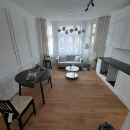 SAMBUD Oleksandr Subbotenko - Jasne wnętrze mieszkania po remoncie: drewniana podłoga, białe ściany z dekoracyjnymi listwami, nowoczesne oświetlenie i meble w neutralnych kolorach. Widok z perspektywy.