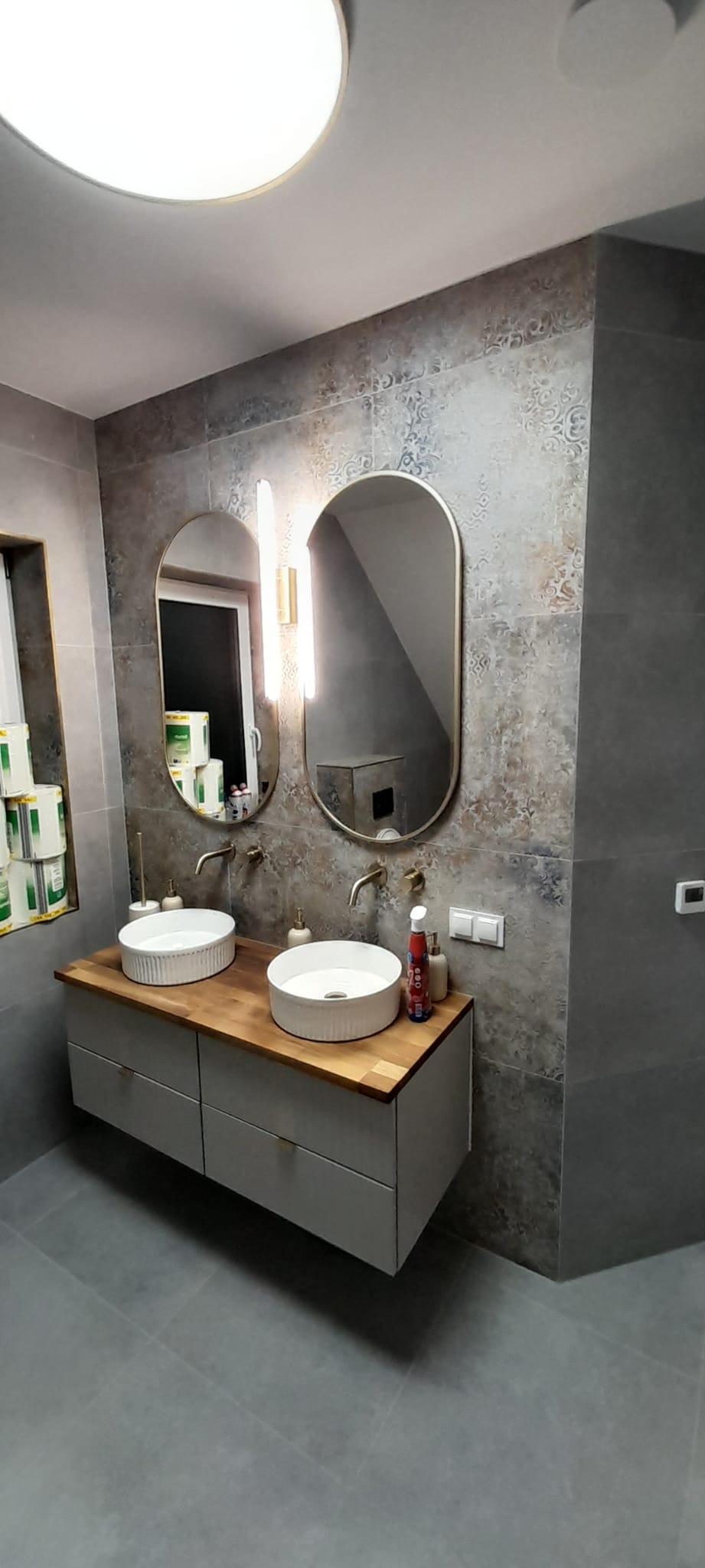 Nowoczesna łazienka z dwoma owalnymi lustrami w złotych ramach, drewnianym blatem i białymi umywalkami nablatowymi. Szare płytki z wzorem na ścianie.