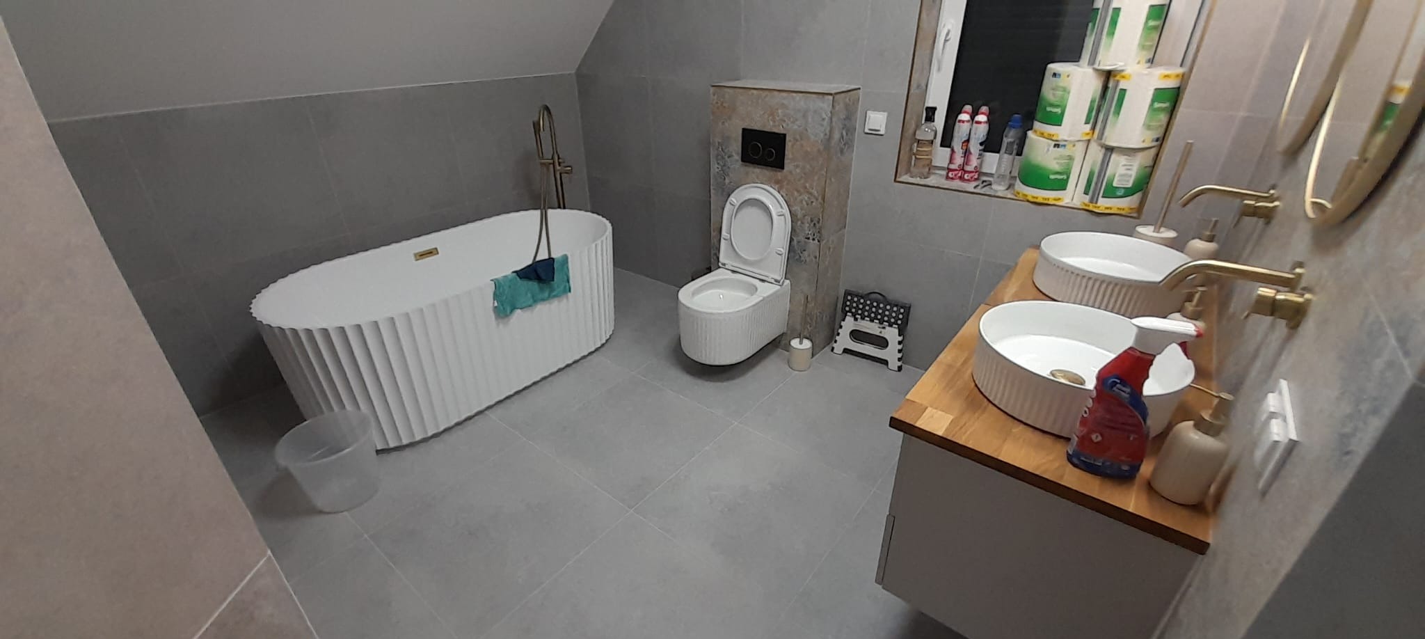 Nowoczesna łazienka w szarościach z wolnostojącą wanną z dekoracyjnym wykończeniem, podwójną umywalką na drewnianym blacie i toaletą w zabudowie.