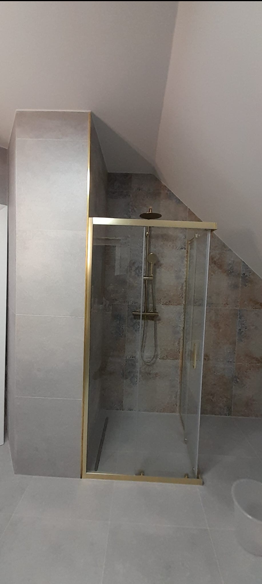 Wykończona kabina prysznicowa z złotymi profilami w łazience na poddaszu, wyłożonej szarymi i beżowymi płytkami. Widoczny zestaw prysznicowy.