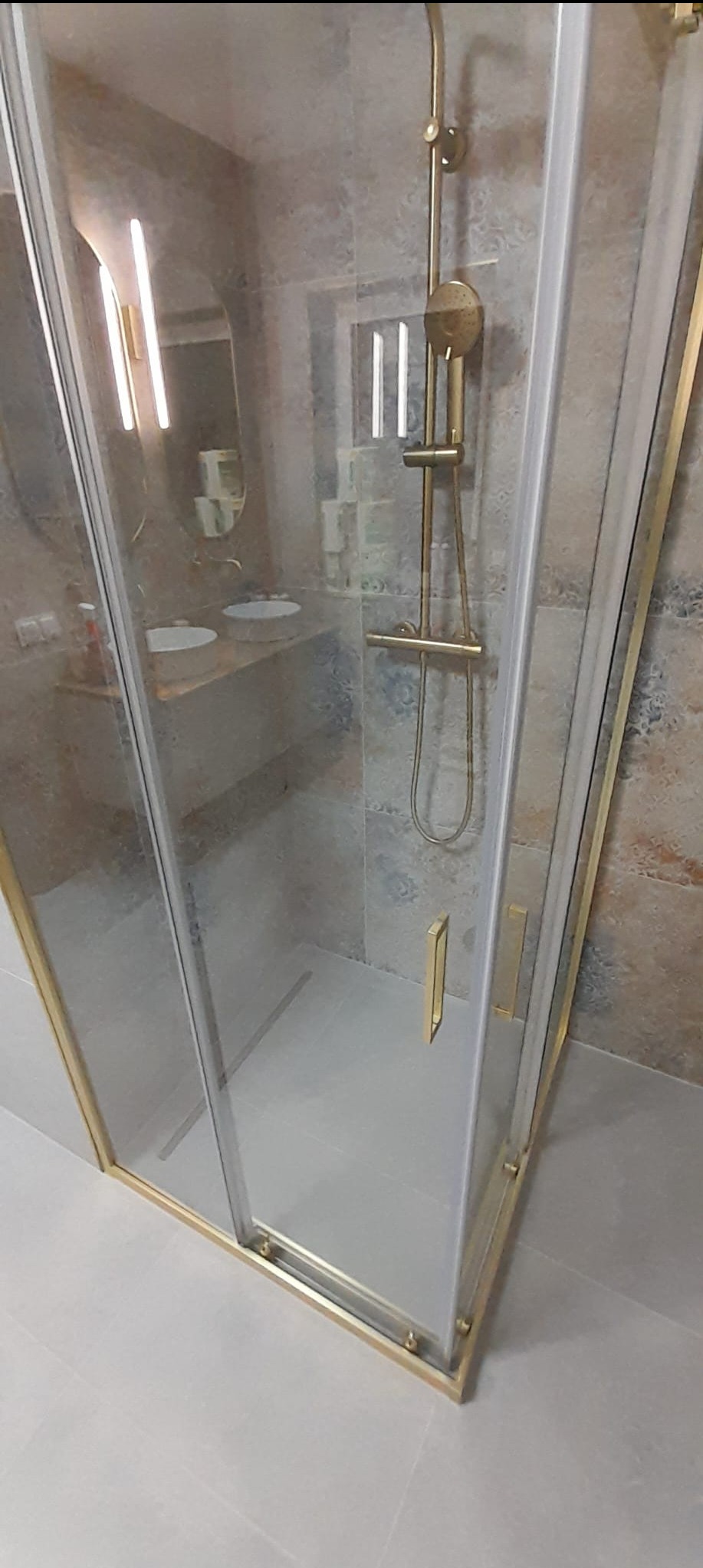 Nowoczesna kabina prysznicowa z złotymi elementami wykończeniowymi, widoczna armatura i płytki w tle. Minimalistyczny design łazienki.