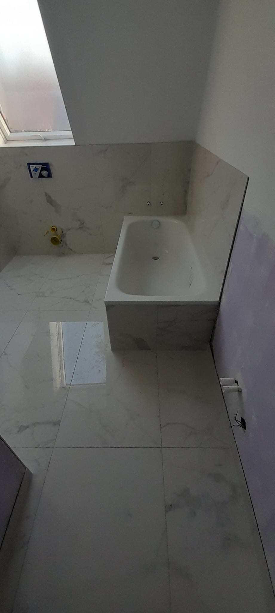 Biała wanna obudowana marmurowymi płytkami w łazience z oknem. Podłoga i ściany wyłożone płytkami imitującymi marmur. Częściowo wykończona ściana z płytą gipsową.