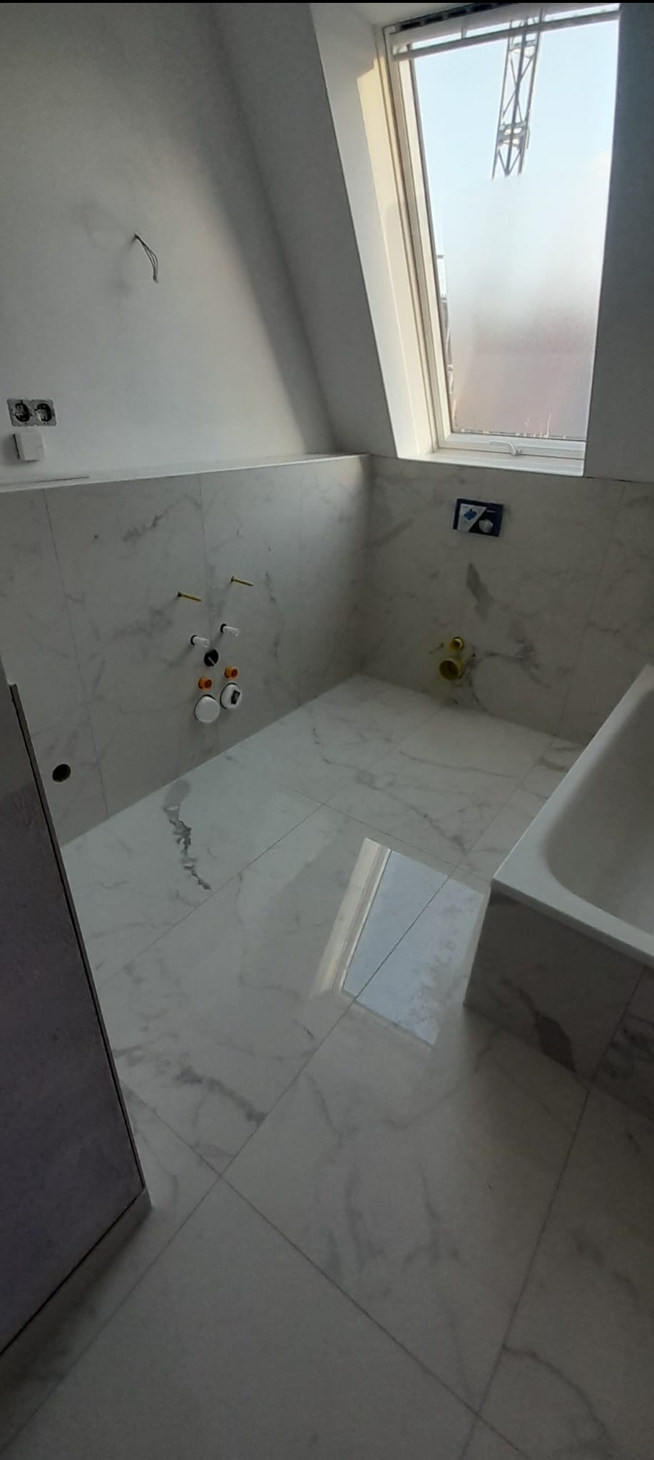 Łazienka na poddaszu w trakcie remontu: białe marmurowe płytki na ścianach i podłodze, widoczne instalacje wodno-kanalizacyjne, okno dachowe. Etap prac przed montażem armatury.