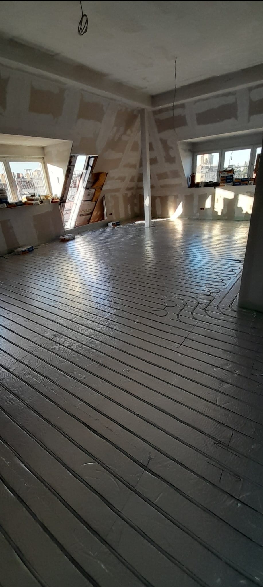 Podłogówka w szarej folii aluminiowej, ułożona w pokoju na poddaszu z oknami i ścianami przygotowanymi pod płytę karton gips. Widoczne materiały budowlane.