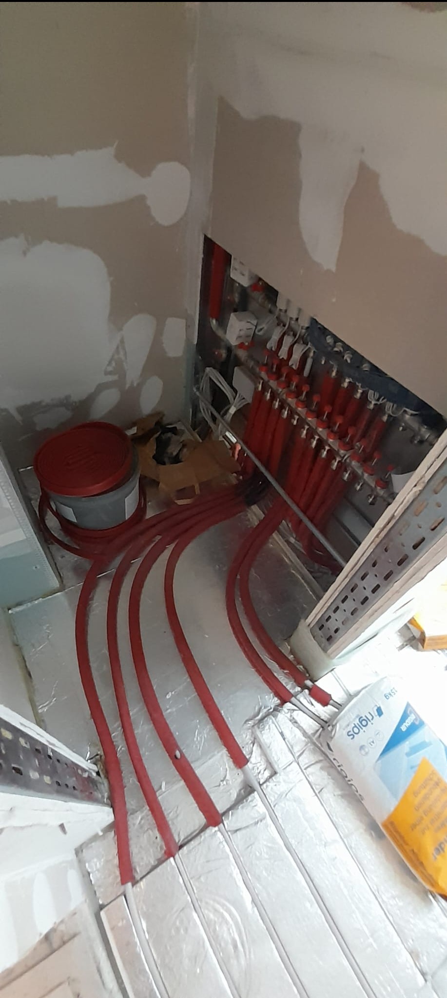 Instalacja ogrzewania podłogowego: czerwone rury rozprowadzone na srebrnej folii izolacyjnej w pomieszczeniu w trakcie wykańczania, widoczny rozdzielacz i puszka z gipsem.