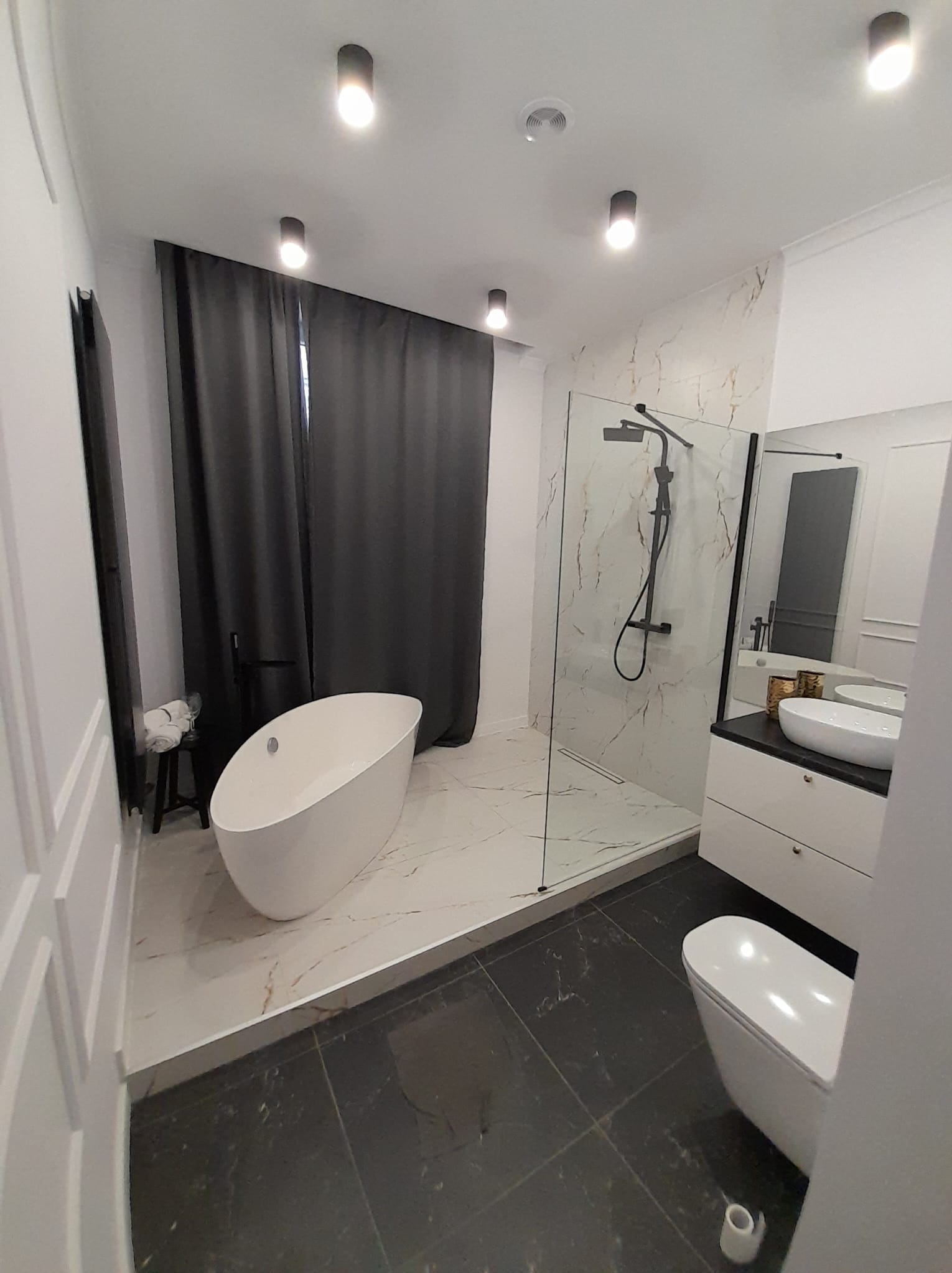 Nowoczesna łazienka z wolnostojącą wanną, kabiną prysznicową typu walk-in i umywalką nablatową. Czarno-biała kolorystyka, marmurowe płytki i minimalistyczne oświetlenie.