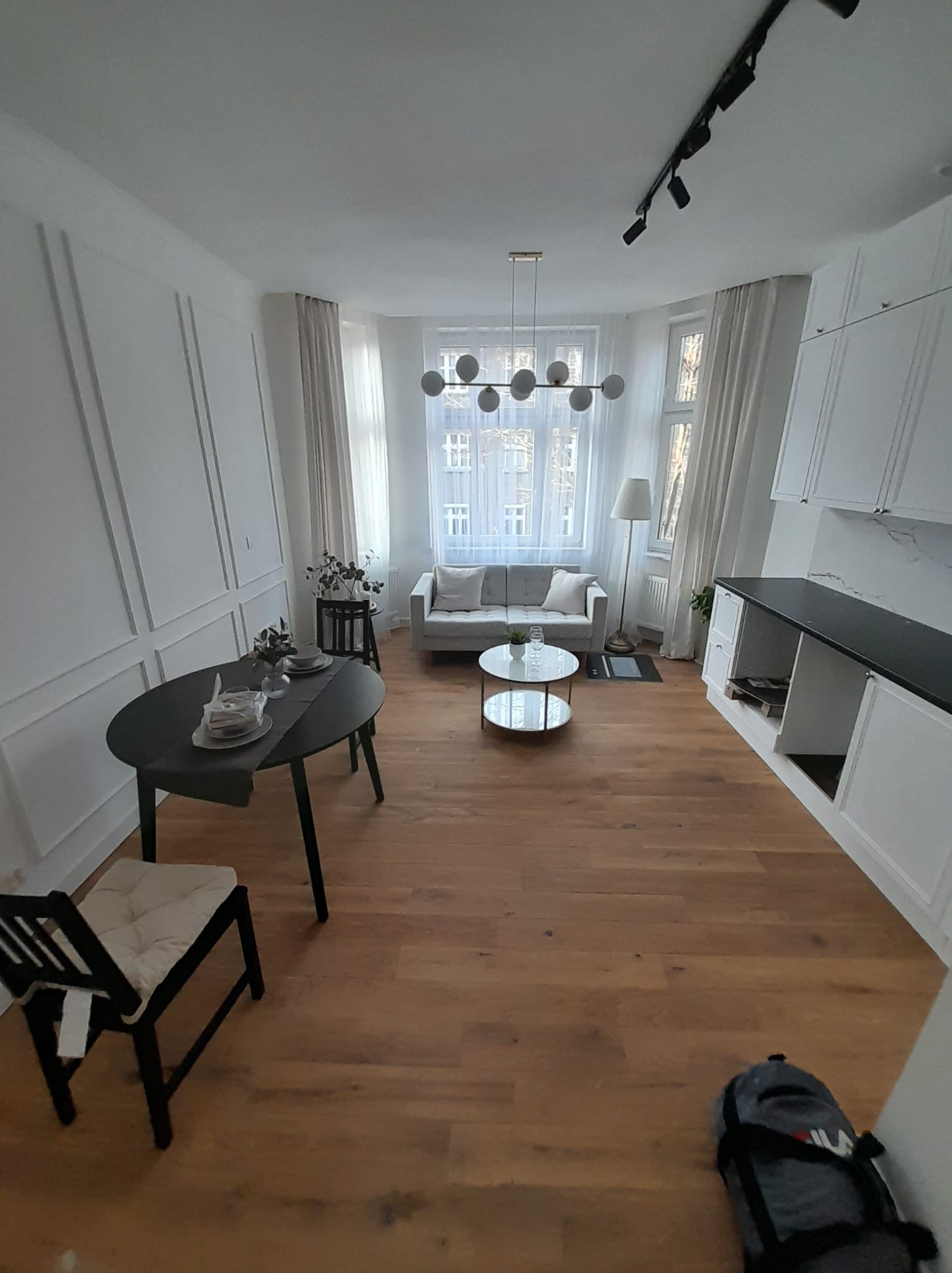 Jasne wnętrze mieszkania po remoncie: drewniana podłoga, białe ściany z dekoracyjnymi listwami, nowoczesne oświetlenie i meble w neutralnych kolorach. Widok z perspektywy.