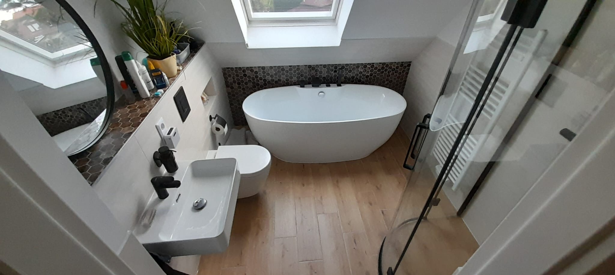 Nowoczesna łazienka na poddaszu w Kostrzynie nad Odrą. Wolnostojąca wanna, umywalka nablatowa z czarną armaturą, drewnopodobne płytki i okrągłe lustro.