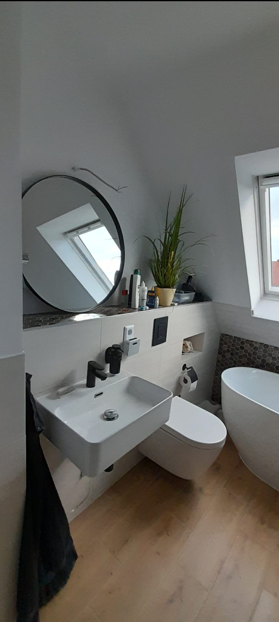 Nowoczesna łazienka na poddaszu z okrągłym lustrem, białą armaturą, drewnianą podłogą i rośliną doniczkową. Uwagę przykuwa nietypowy kształt umywalki i mozaika na ścianie.