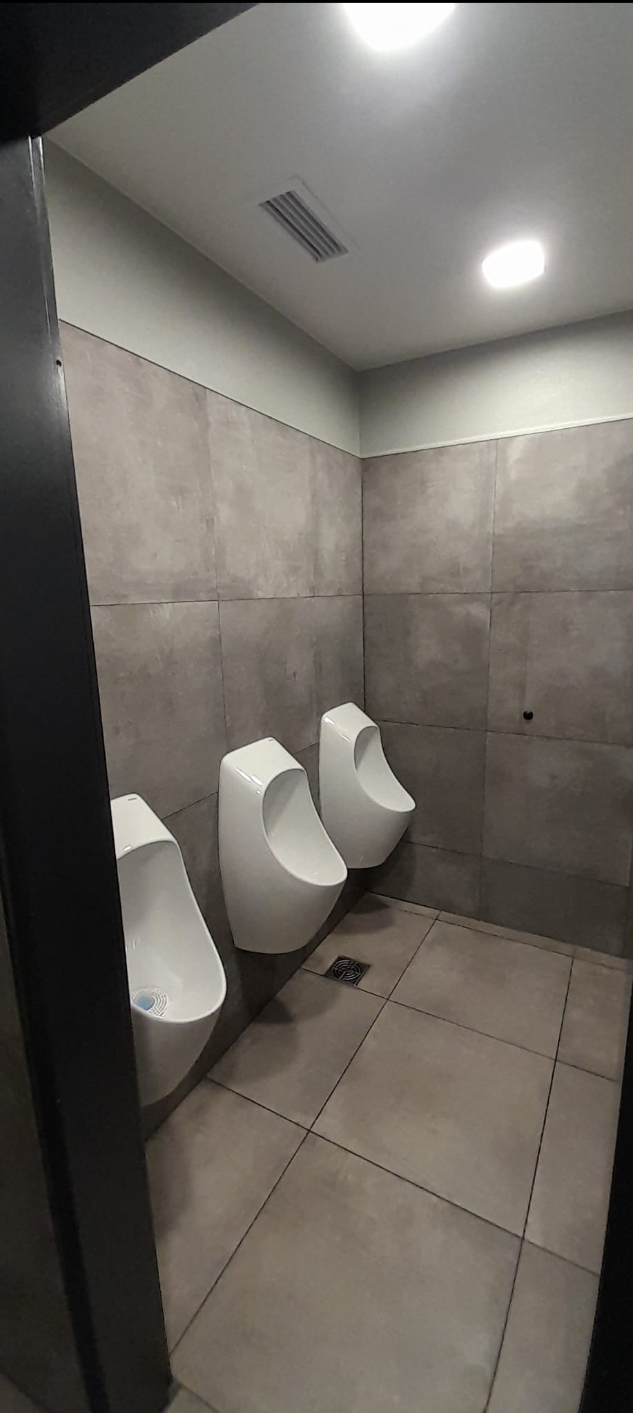 Nowoczesna toaleta publiczna z trzema białymi pisuarami na szarej, kwadratowej płytce. Minimalistyczny design, surowy styl, widoczny fragment czarnej ramy drzwi.