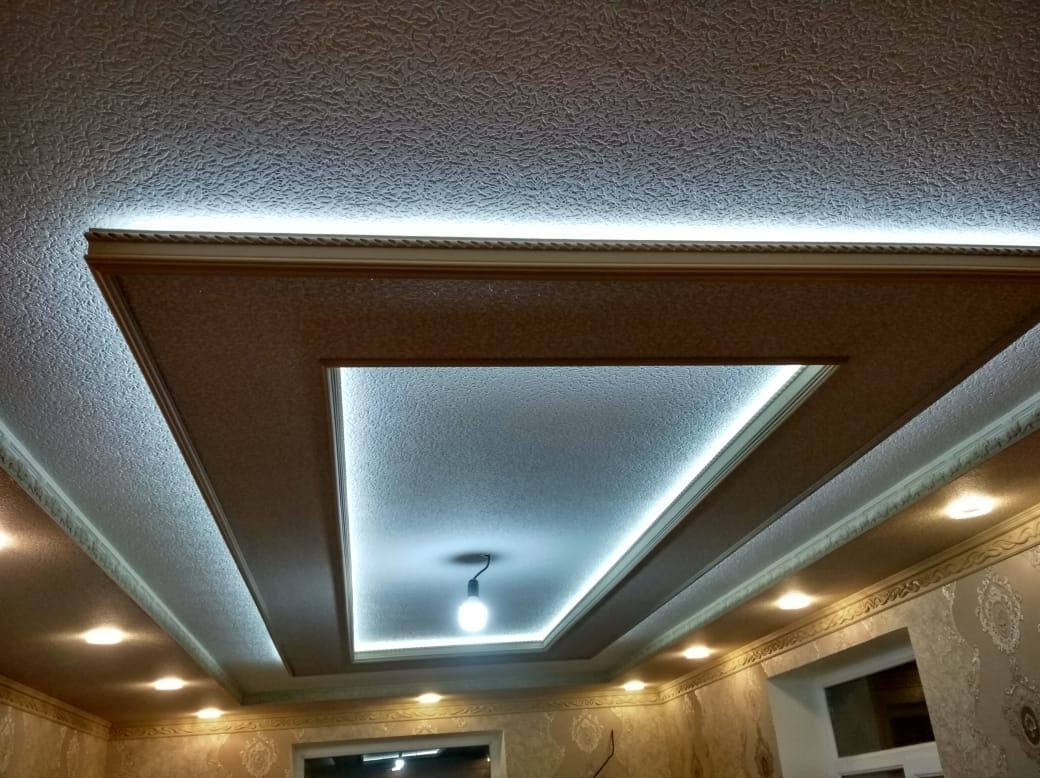 Dekoracyjny sufit podwieszany z oświetleniem LED, widok z dołu. Geometryczny wzór z ramą i centralnym punktem świetlnym. Beżowe ściany z wzorem w tle.