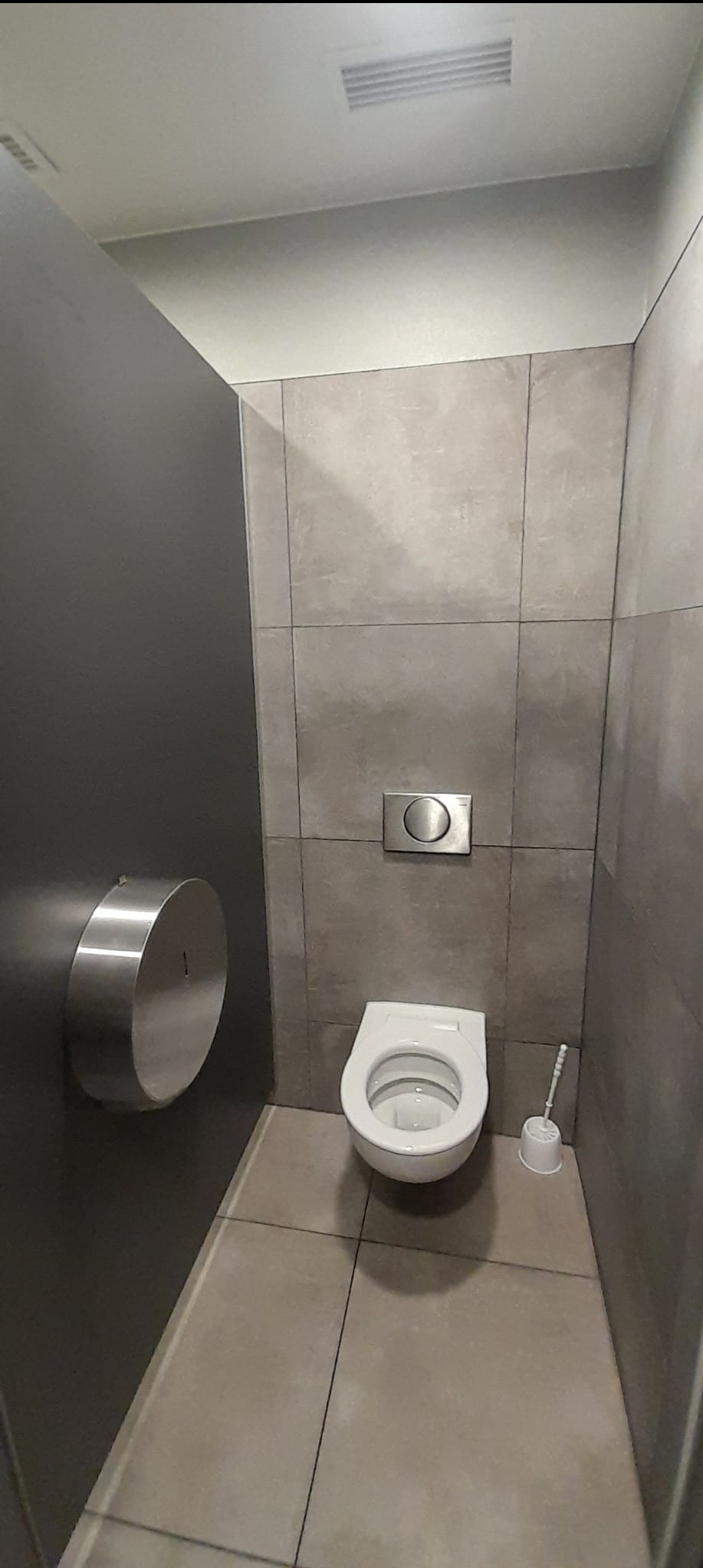 Nowoczesna toaleta w szarościach z podtynkową spłuczką i szczotką WC. Minimalistyczny design, płytki imitujące beton, metalowy podajnik na papier toaletowy.