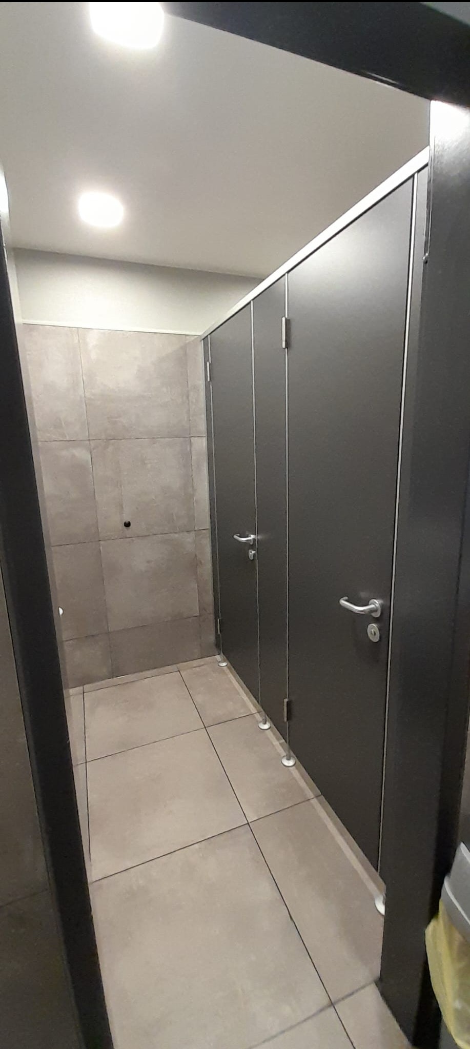 Wykończona toaleta publiczna z kabinami w odcieniach szarości i beżu. Widoczne płytki na ścianach i podłodze, metalowe ramy kabin i uchwyty drzwiowe.