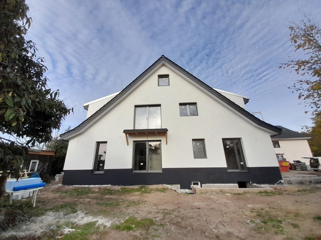 Dwukondygnacyjny dom z ocieploną i świeżo pomalowaną elewacją w kolorze białym z czarnym cokołem, widoczne okna z szarymi ramami, teren wokół domu w trakcie prac wykończeniowych.