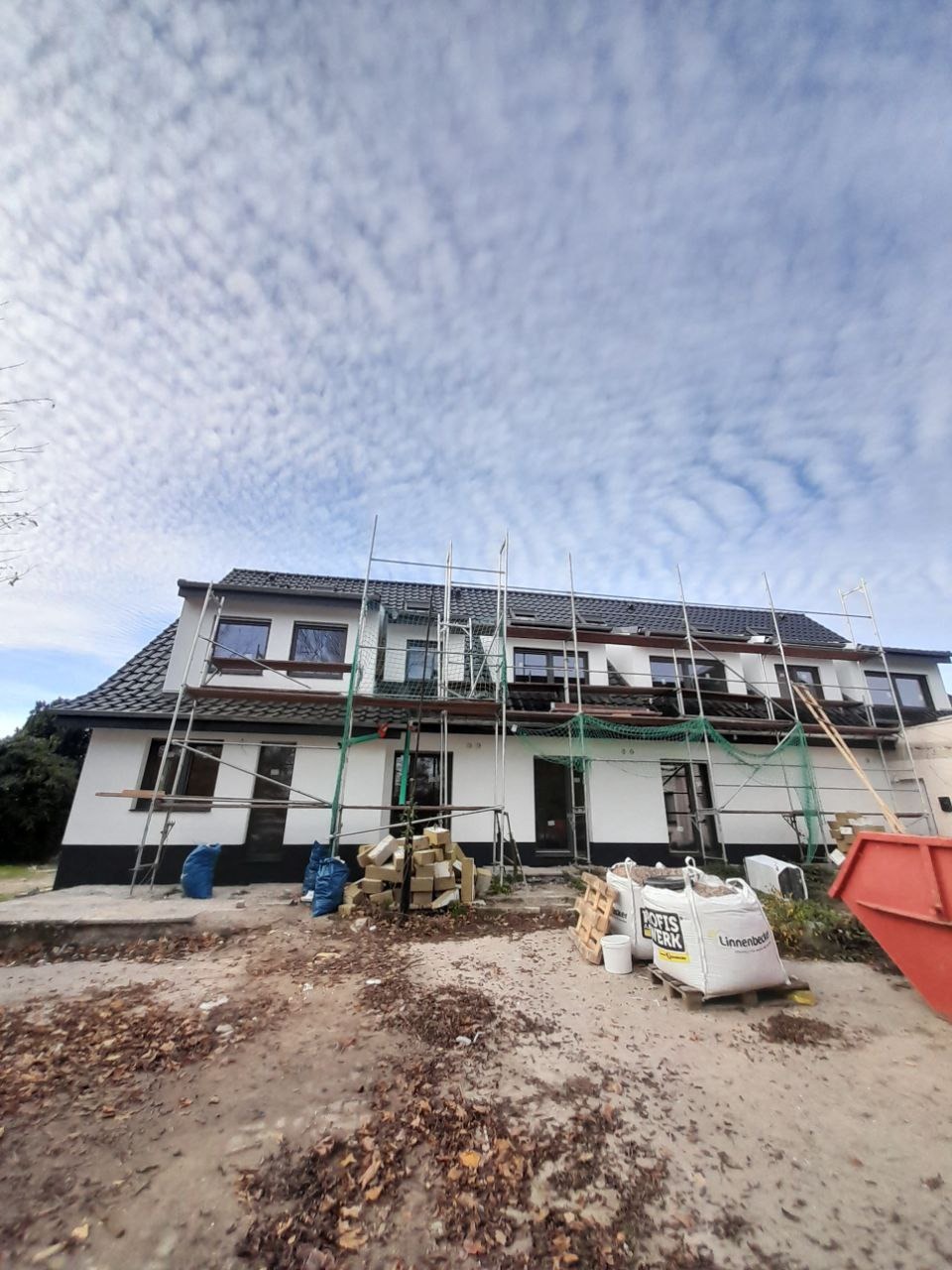 Dwupiętrowy dom w trakcie ocieplania elewacji, otoczony rusztowaniami i materiałami budowlanymi, z workami zaprawy i gruzem na pierwszym planie, sfotografowany w pochmurny dzień.