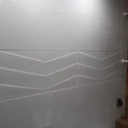 Ściana w łazience wyłożona białymi, poziomymi płytkami z delikatnym, geometrycznym wzorem 3D, oświetlona górnym światłem.