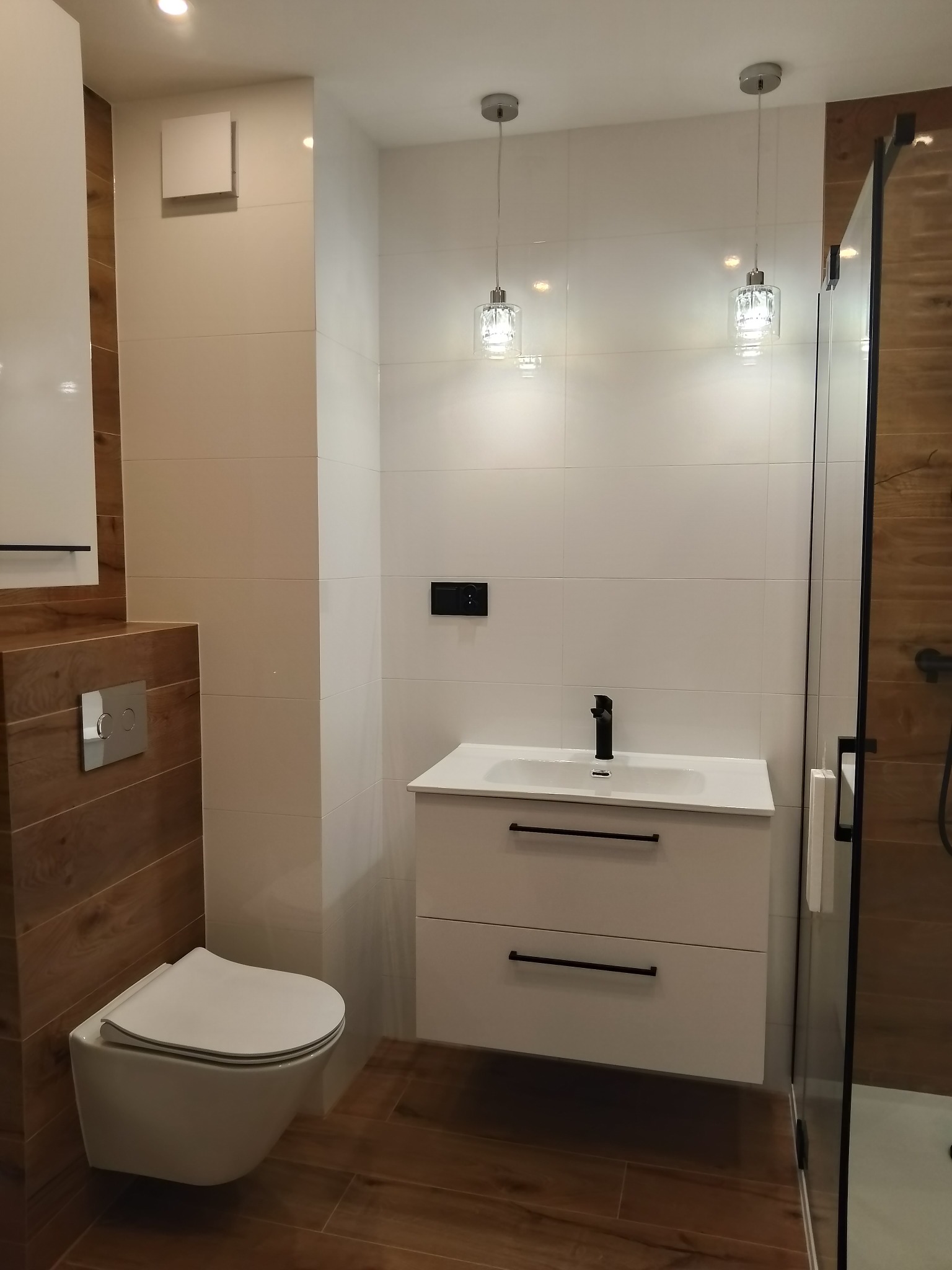 Nowoczesna łazienka z podwieszaną toaletą, białą umywalką z szafką i kabiną prysznicową z drewnopodobnymi płytkami, oświetlona wiszącymi lampami z kryształkami.