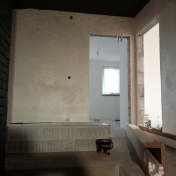 Malowanie i Szpachlowanie - Ściana z nałożoną dekoracyjną gładzią w odcieniach beżu i szarości, widoczne puszki farb i materiały wykończeniowe, wnętrze w trakcie remontu z surowymi framugami drzwi.