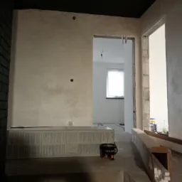 Ściana z nałożoną dekoracyjną gładzią w odcieniach beżu i szarości, widoczne puszki farb i materiały wykończeniowe, wnętrze w trakcie remontu z surowymi framugami drzwi.