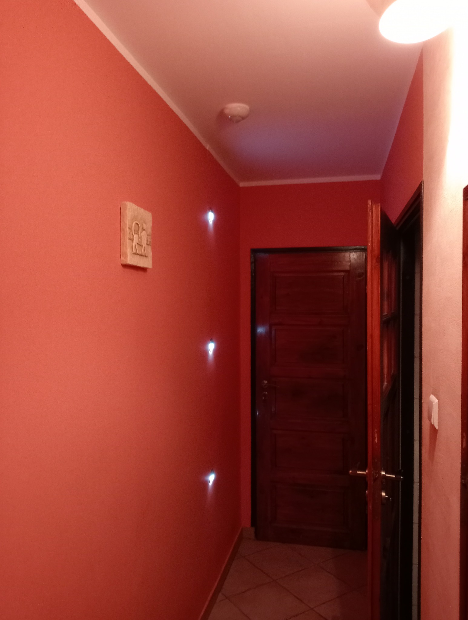 Korytarz pomalowany na koralowo-czerwony kolor z drewnianymi drzwiami i dekoracyjnym oświetleniem punktowym w ścianie.